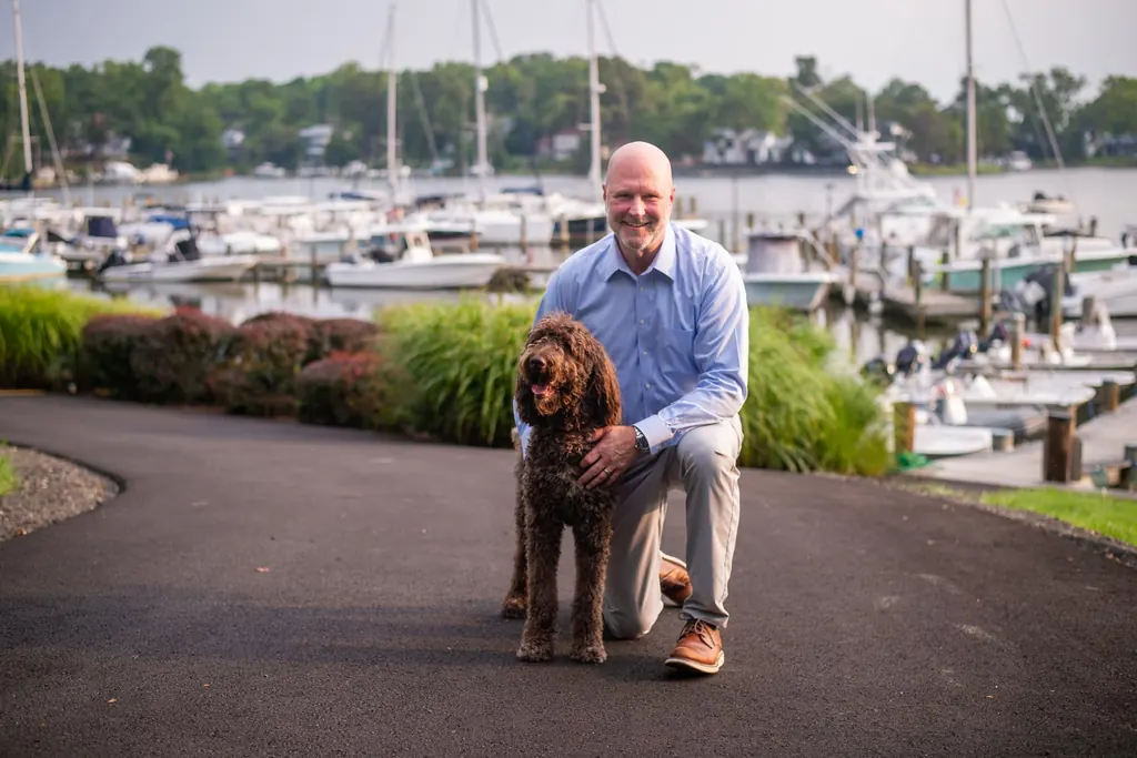 Jason C. Johnson kneeling with his dog on a path beside an Annapolis marina